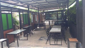 An outdoor covered seating area with tables and chairs, surrounded by greenery. - Lilly Home (Khaem Son)