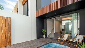 A modern outdoor swimming pool with wooden decking, a glass door leading to a staircase, and lounge chairs. - Peaceful 2BR Umalas Villa Near Seminyak (Badung)