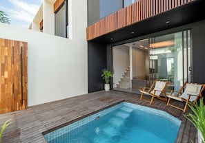 Ein moderner Außenpool mit Holzterrasse, eine Glastür zu einer Treppe und Liegestühle.