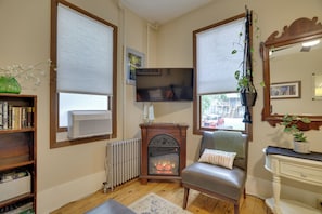A living room with a fireplace, a television, a bookshelf, and a window with blinds. - Walk to Main St Stroudsburg! Historic Dtwn Home (Stroudsburg)