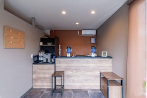 Zona de recepción con mostrador, dos taburetes de madera, un tablón de anuncios, una máquina de café y un aire acondicionado instalado en la pared.