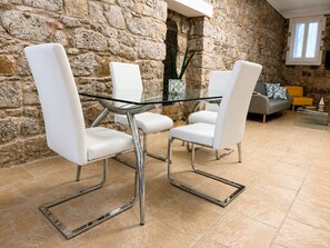 A dining area with a glass table and white chairs, a stone wall, and a sofa with cushions. - Maison Tabarka: Comfort and Design Suites in the Historic Center of Carloforte (Carloforte)