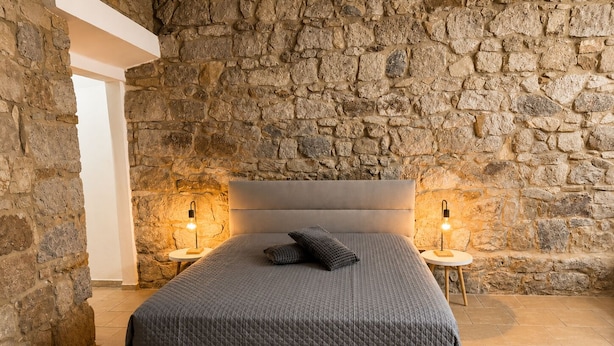 A bedroom with a stone wall, a bed with a grey quilt, two bedside lamps, and a white ceiling structure. - Maison Tabarka: Comfort and Design Suites in the Historic Center of Carloforte (Carloforte)