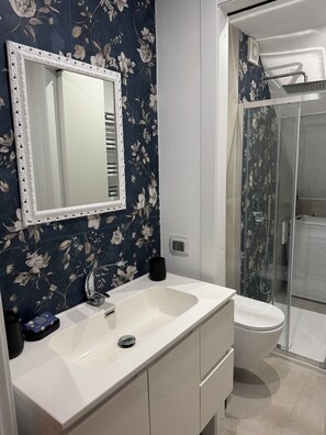 A bathroom with a white sink, a mirror with a decorative frame, a black mug, and a shower area with a glass door. - Maison Tabarka: Comfort and Design Suites in the Historic Center of Carloforte (Carloforte)