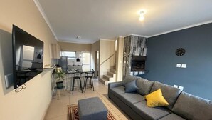 Uma sala de estar moderna com um sofá cinzento, uma mesa de centro e um tapete estampado. Há uma área de cozinha com uma mesa de jantar e cadeiras, e uma escada que leva a outro andar.