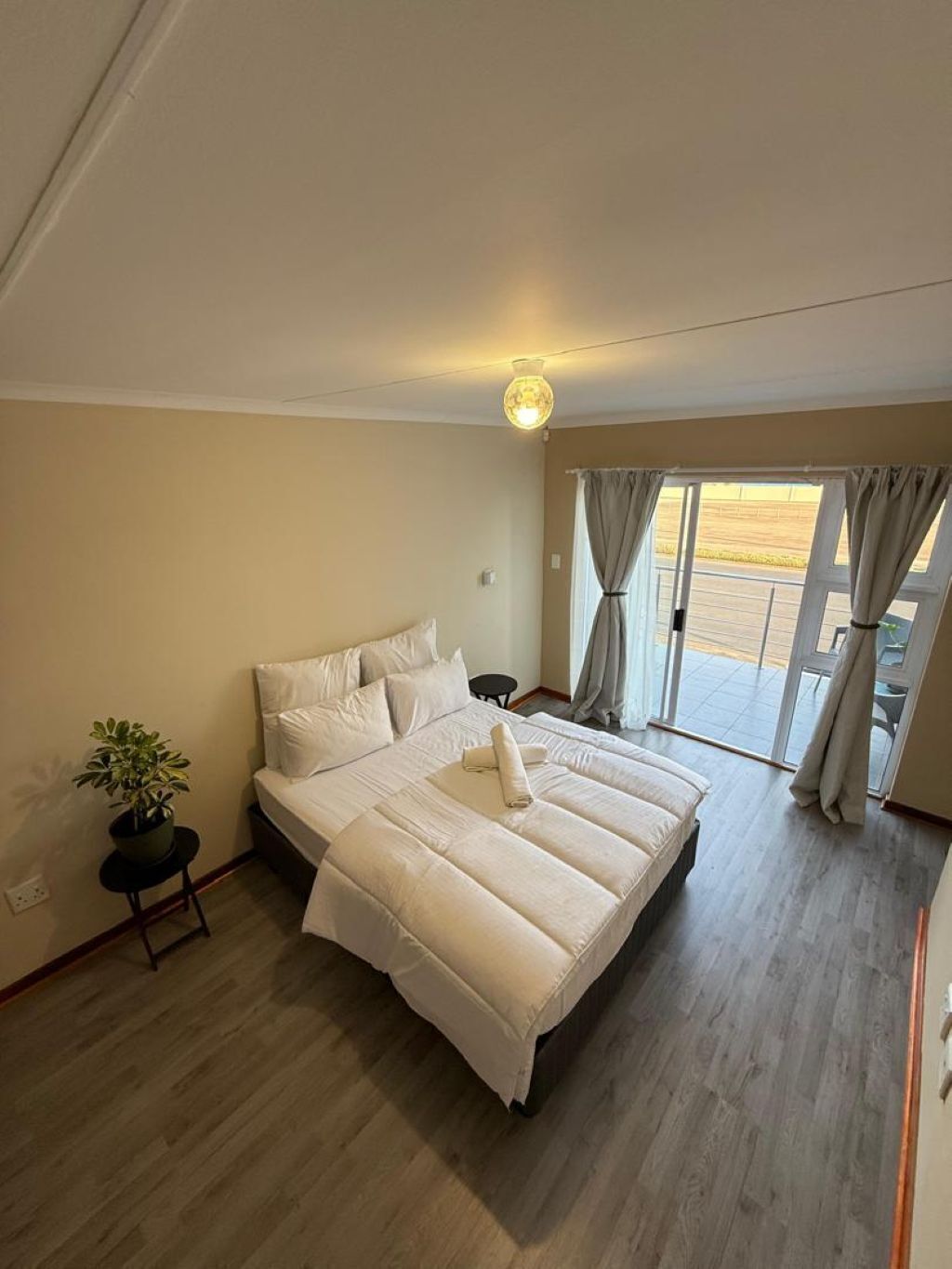 A bedroom with a large bed, a potted plant, and a view through a sliding glass door.