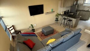 Uma sala de estar moderna com um sofá modular cinza, uma televisão de tela plana na parede e uma pequena área de cozinha com banquetas.
