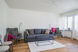 A living room with a grey sofa, striped chairs, a white rug, and a window with curtains.