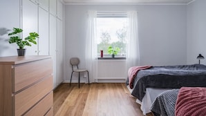 A bedroom with a wooden dresser, a chair, a window with curtains, a radiator, and a bed with a blanket.