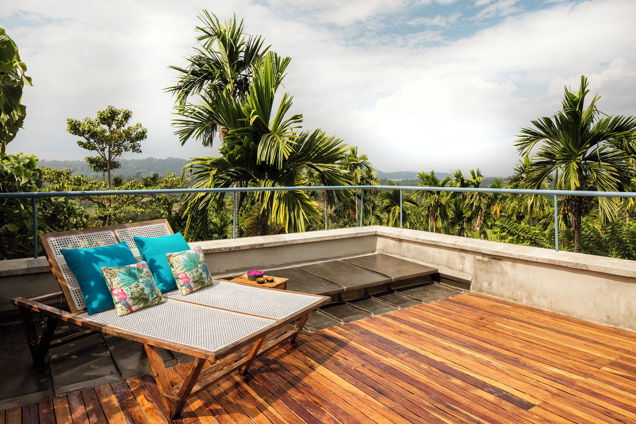 A wooden deck with a lounge chair and cushions, overlooking a lush landscape with palm trees.