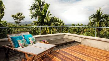 A wooden deck with a lounge chair and cushions, overlooking a lush landscape with palm trees.