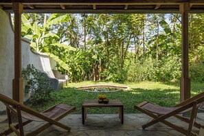 A covered outdoor seating area with two wooden chairs and a table, surrounded by lush greenery and a small pond.