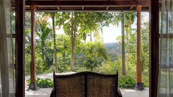 A wooden bench with a woven backrest under a thatched roof, overlooking a lush garden with tall trees and a hilly landscape.