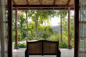 A wooden bench with a woven backrest under a thatched roof, overlooking a lush garden with tall trees and a hilly landscape.