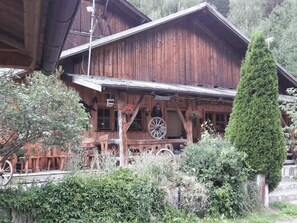 Un edificio de madera con un porche, una rueda y un árbol alto.