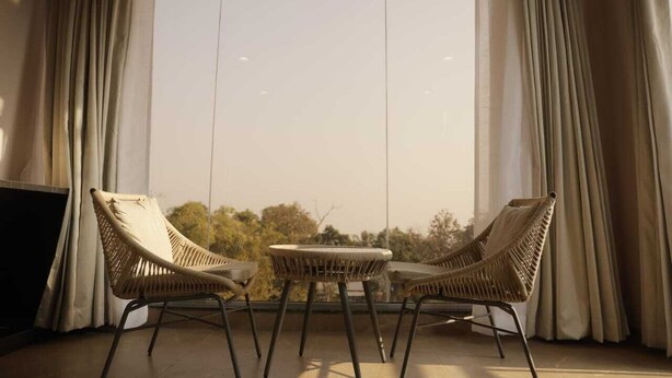 A room with a large window, two wicker chairs, a small table, and a television on a stand. - Shourya Resort and lawn Panhala (Panhala)