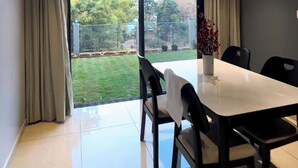 A dining area with a table and chairs, a ceiling fan, and a view of a garden through large windows. - Shourya Resort and lawn Panhala (Panhala)