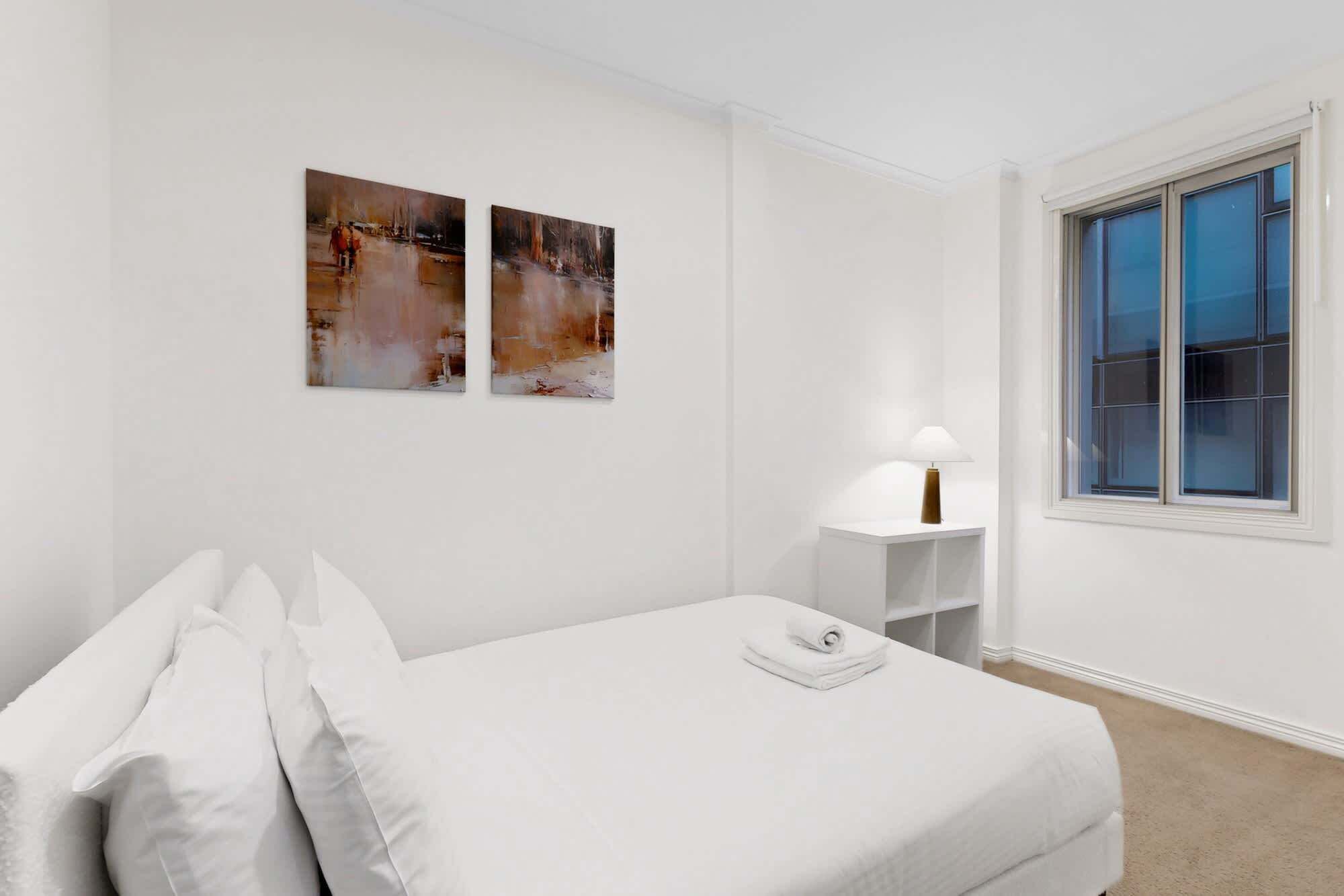 A hotel room with a bed, two paintings on the wall, a bedside table with a lamp, and a window with a view.
