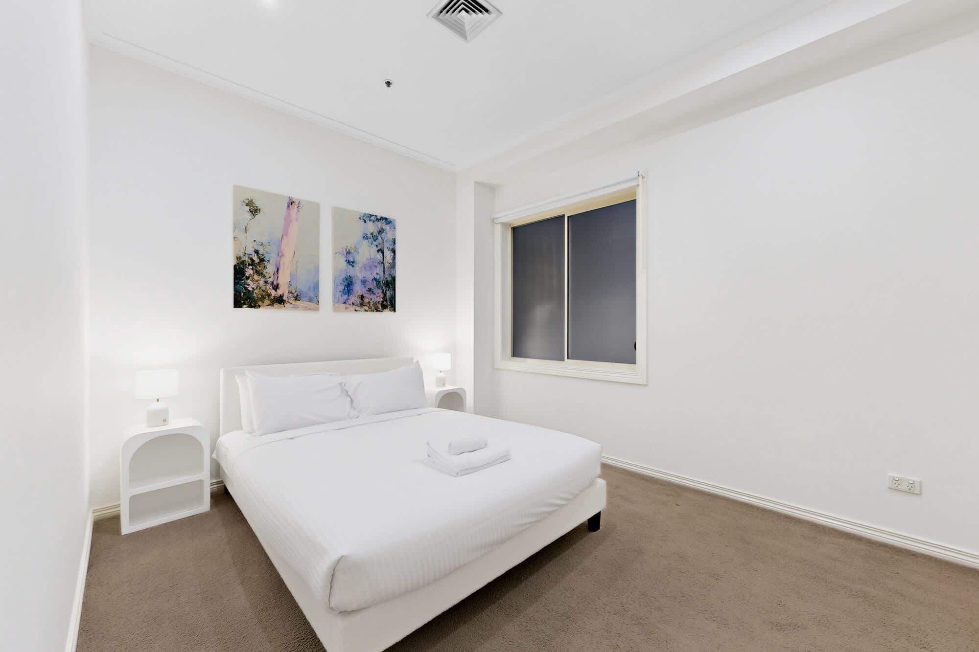 A bedroom with a bed, two bedside tables, a window, and two wall-mounted paintings.