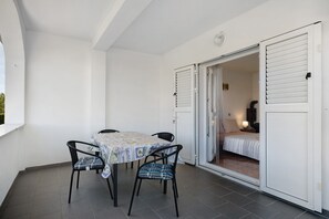 Ein kleiner Balkon mit Tisch und Stühlen, eine Schiebetür, die zu einem Schlafzimmer führt, und eine weiße Außenwand.