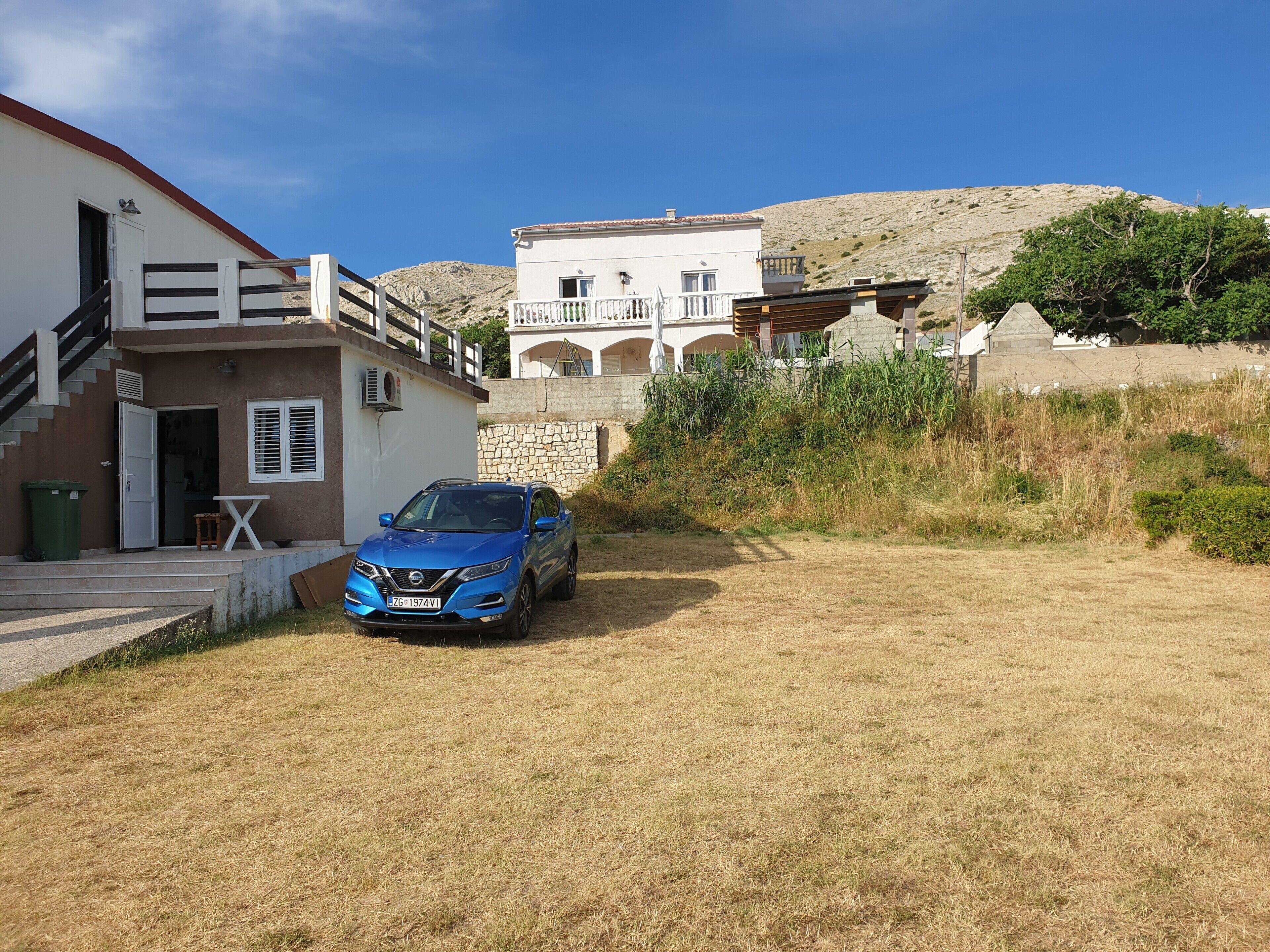 A residential area with a blue car parked in front of a house with a stone wall and a staircase leading to a balcony.