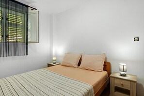 Un dormitorio con cama, dos mesitas de noche, una mesita auxiliar, una ventana con persianas y un reloj de pared.