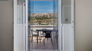 An open door leading to a balcony with a table and chairs, overlooking a hilly landscape with buildings. - Apartment 'Volarić Pag' with Sea View, Balcony and Air Conditioning (Pag)