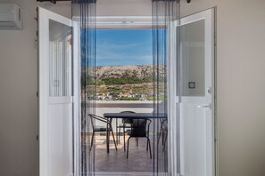 Een open deur die leidt naar een balkon met een tafel en stoelen, met uitzicht op een glooiend landschap met gebouwen.