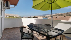 A balcony with a table and chairs, a green umbrella, and a view of a coastal town and mountains. - Apartment 'Volarić Pag' with Sea View, Balcony and Air Conditioning (Pag)