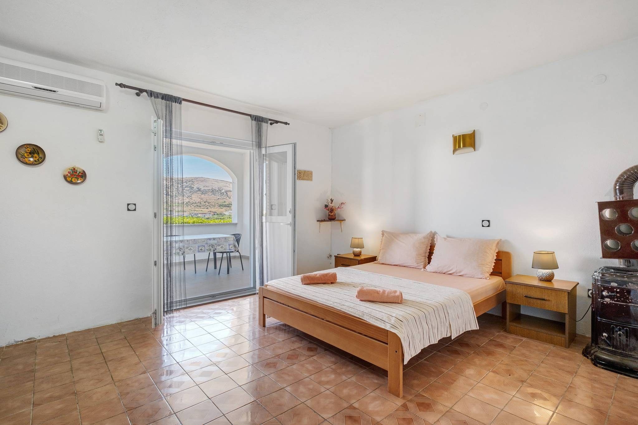 A bedroom with a bed, bedside tables, a lamp, a heater, and a view through a glass door.