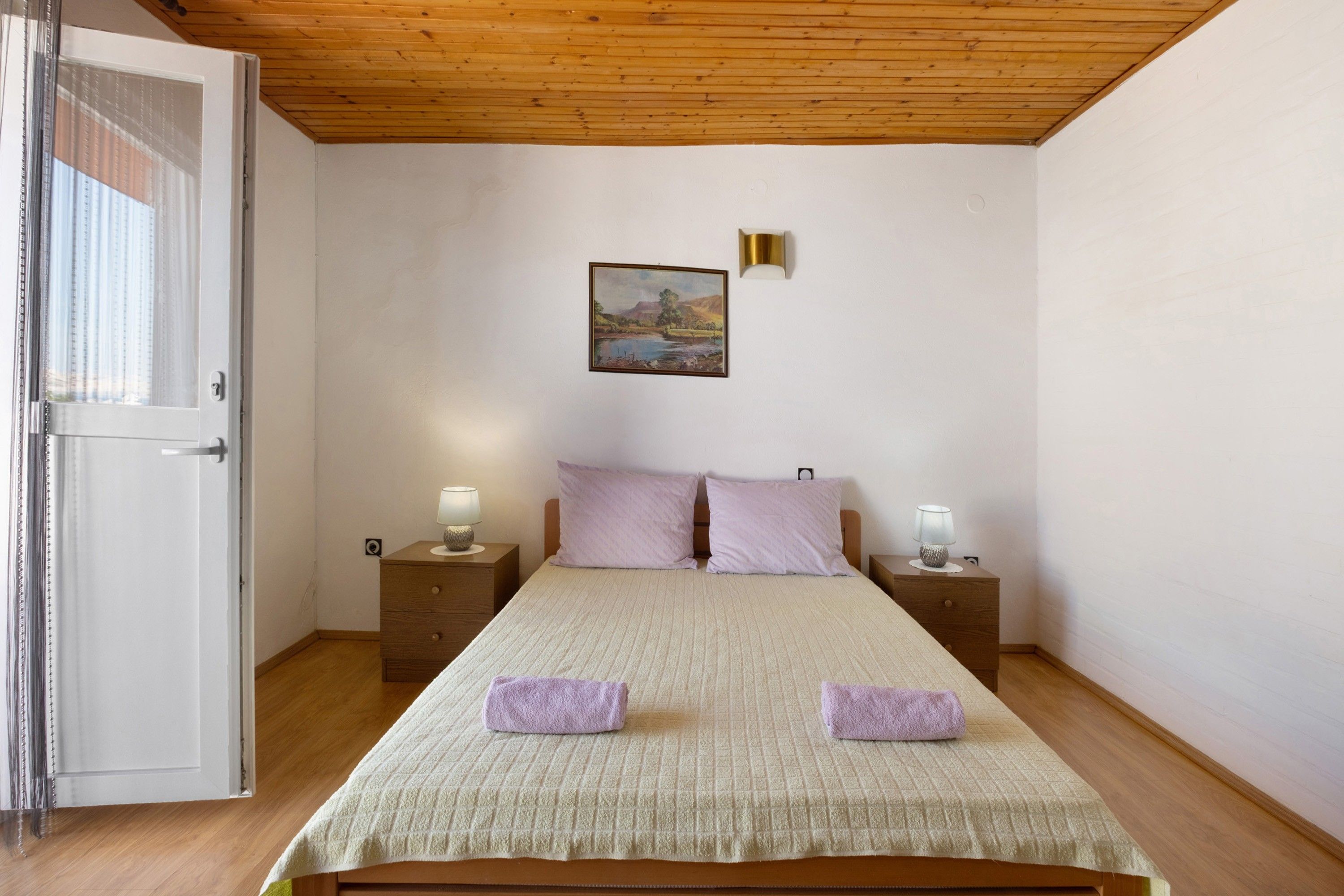 A bedroom with a wooden ceiling, a bed with a quilted bedspread, two nightstands with lamps, and a painting on the wall.