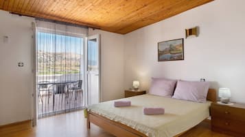 A bedroom with a wooden ceiling, a bed with a white bedspread, two bedside lamps, a painting on the wall, and a view through a window with a curtain.