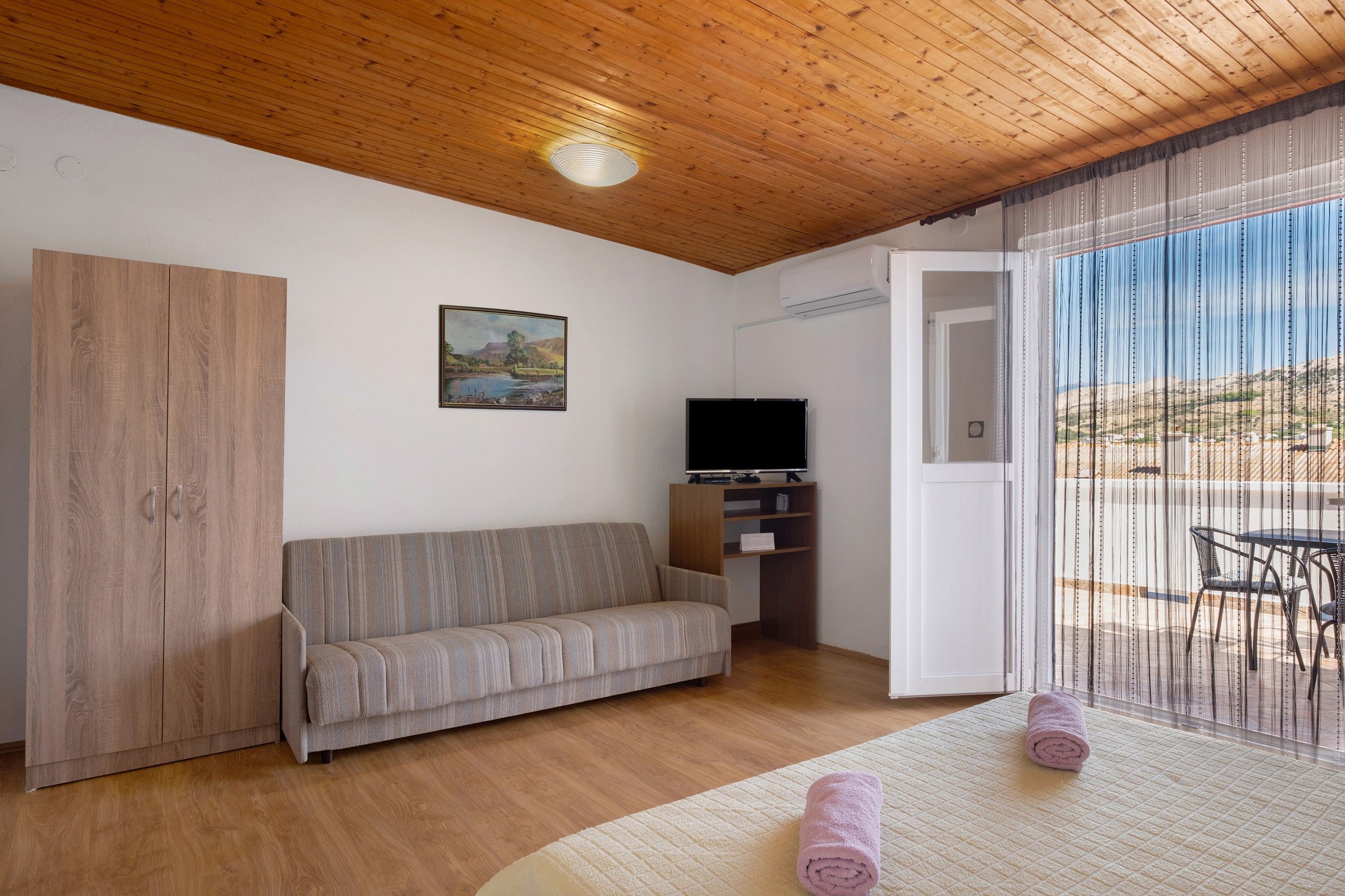 A room with a wooden ceiling, a sofa, a television, and a view through a window with curtains.