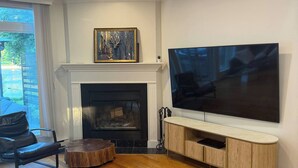 Uma sala de estar com lareira, uma televisão de tela plana grande fixada em um console de madeira, um sofá de couro e uma mesa de jantar.
