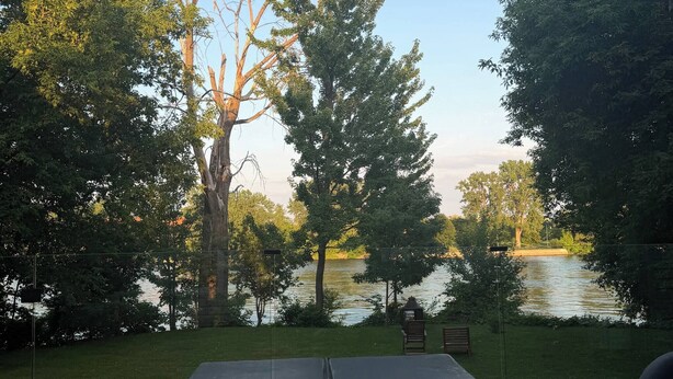 Um terraço com mesa e cadeiras, com vista para um lago com árvores e um banco.