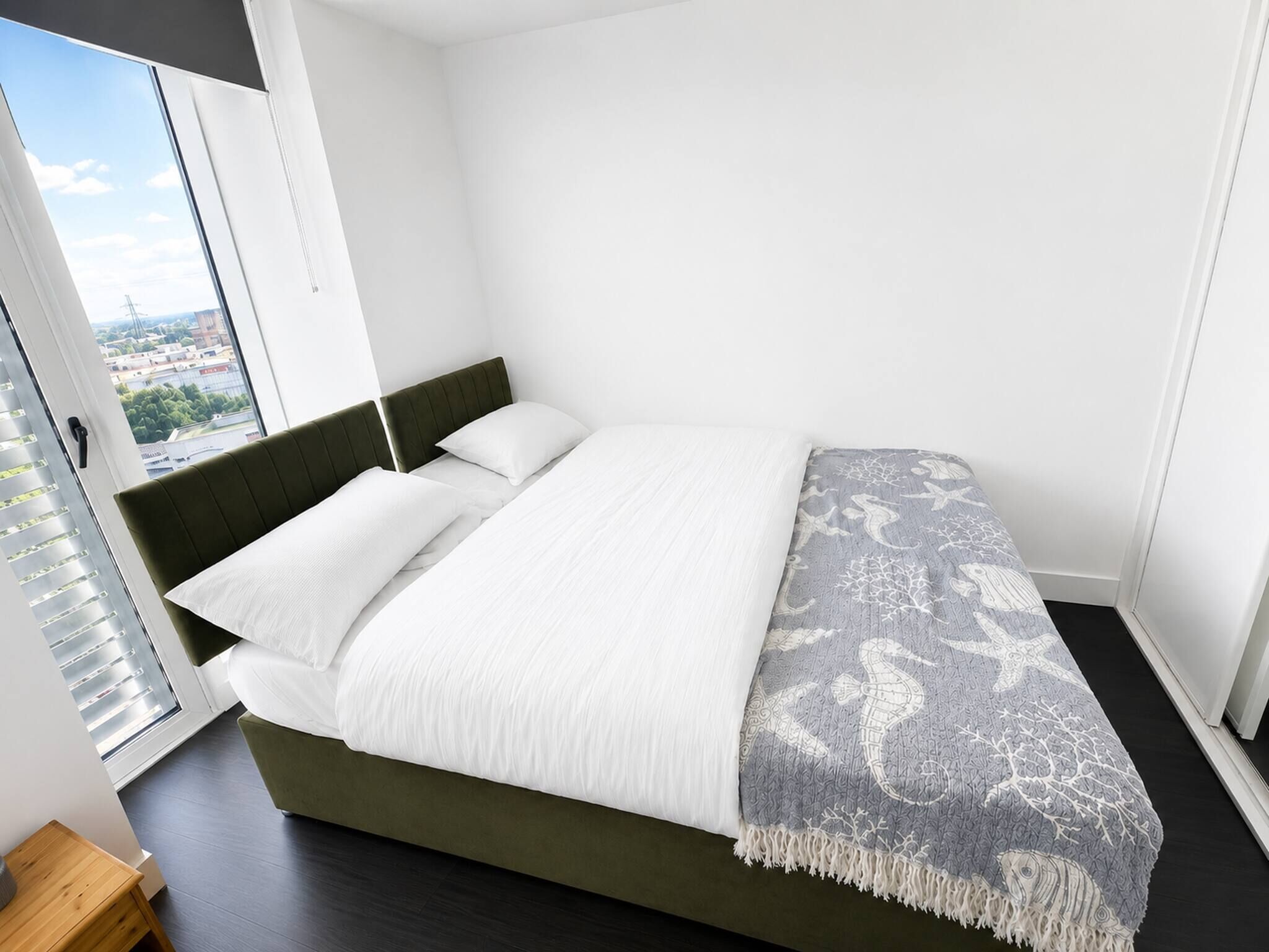Un dormitorio moderno con una cama, ropa de cama blanca, una manta estampada y vistas a través de un gran ventanal.