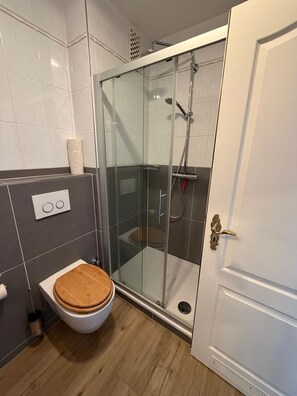A modern bathroom with a glass shower enclosure, a wooden toilet, and a white door. - Strandstraße Building, Apartment 33 (Sylt)