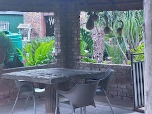 Eine Terrasse im Freien mit einem rustikalen Holztisch und modernen Stühlen, umgeben von üppiger Vegetation und einer Backsteinmauer.