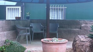 An outdoor patio with a table and chairs under a blue umbrella, a potted plant, and a window with a curtain.
