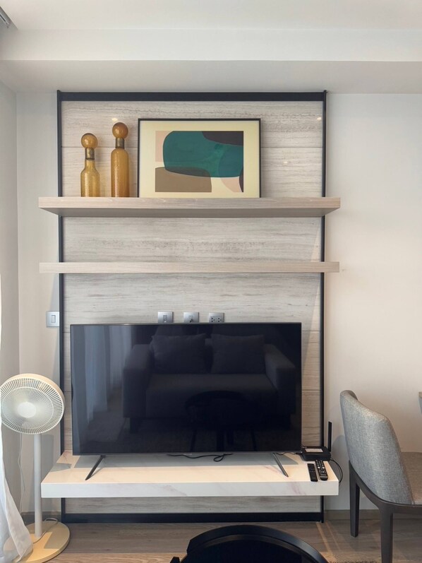Een moderne woonkamer met een grote flatscreen televisie op een witte standaard, een grijze fauteuil en een kleine tafel met afstandsbedieningen.
