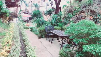 A garden pathway with a bench and table, surrounded by various plants and a building in the background.
