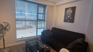 A living room with a black sofa, a white armchair, a glass coffee table, a floor lamp, and a large window with blinds. - Canary wharf view apartment (London)