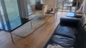 A modern living room with a black leather sofa, a glass partition, a white armchair, and a small table with a laptop. - Canary wharf view apartment (London)