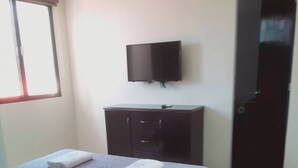 Une chambre à coucher comprenant un lit, un téléviseur à écran plat fixé au mur, une commode de couleur sombre et une porte.