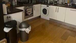 A compact kitchen with white cabinets, a stainless steel range hood, a washing machine, and a trash bin. - Charming 1-Bedroom Apartment in Central London (London)