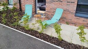 A brick building with a sliding glass door entrance, two outdoor lounge chairs, a small wooden table, and a garden area with mulch and plants. - Holiday home with private bathroom (Edinburgh)