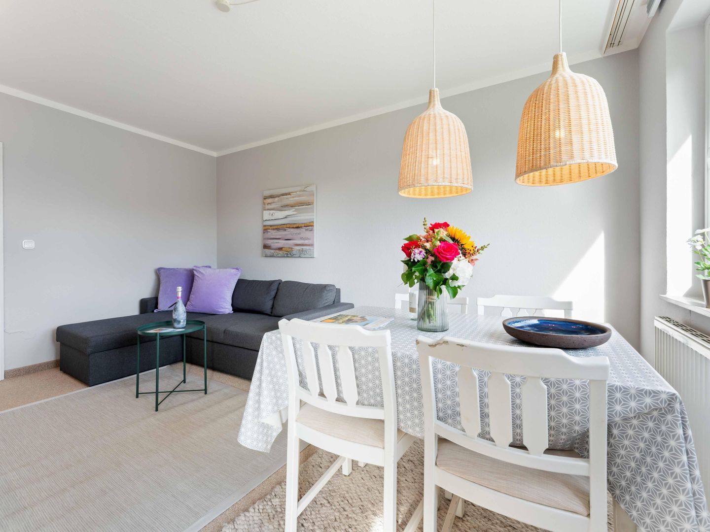 Ein modernes Wohnzimmer mit Esstisch, weißen Stühlen, einem grauen Sofa und zwei Weidenkorb-Pendelleuchten.