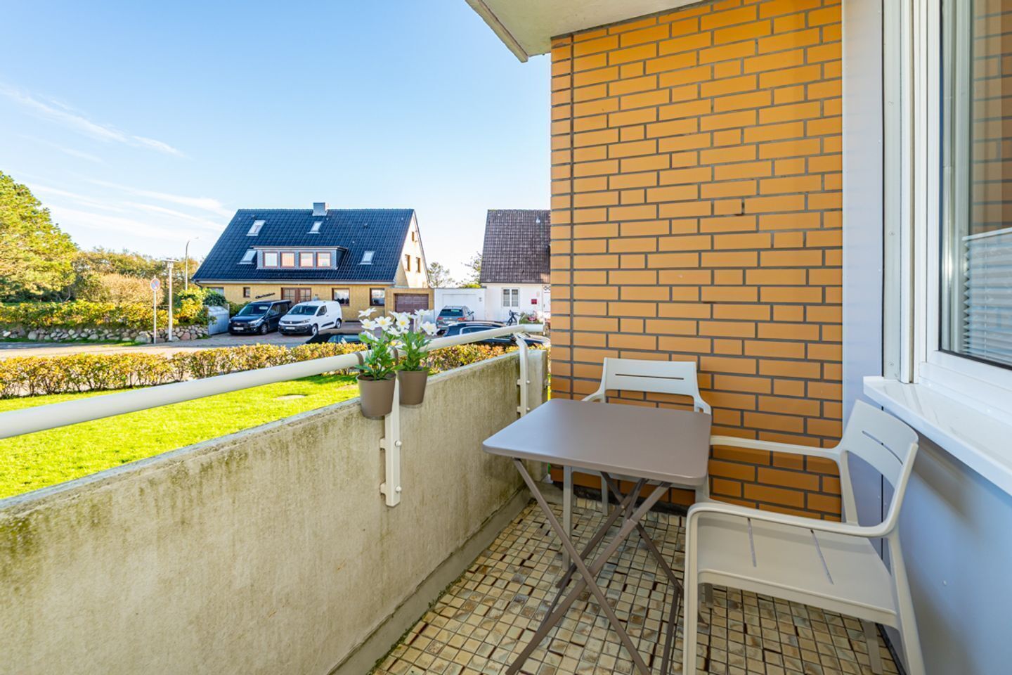 A balcony with a table and chairs, potted plants, and a view of a street with parked cars and houses.
