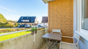 En balkon med et bord og stole, plantekasser og udsigt til en gade med parkerede biler og huse.