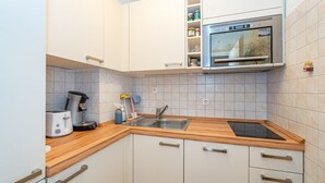 Et kompakt køkken med hvide skabe, en indbygget ovn, kaffemaskine og vask.